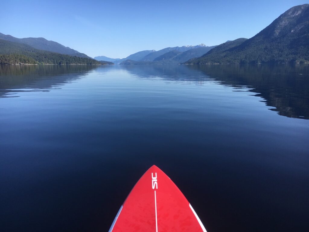 A first person view while standing on a red paddle board looking at the glassy calm waters of the Sechelt Inlet on a sunny day. 
